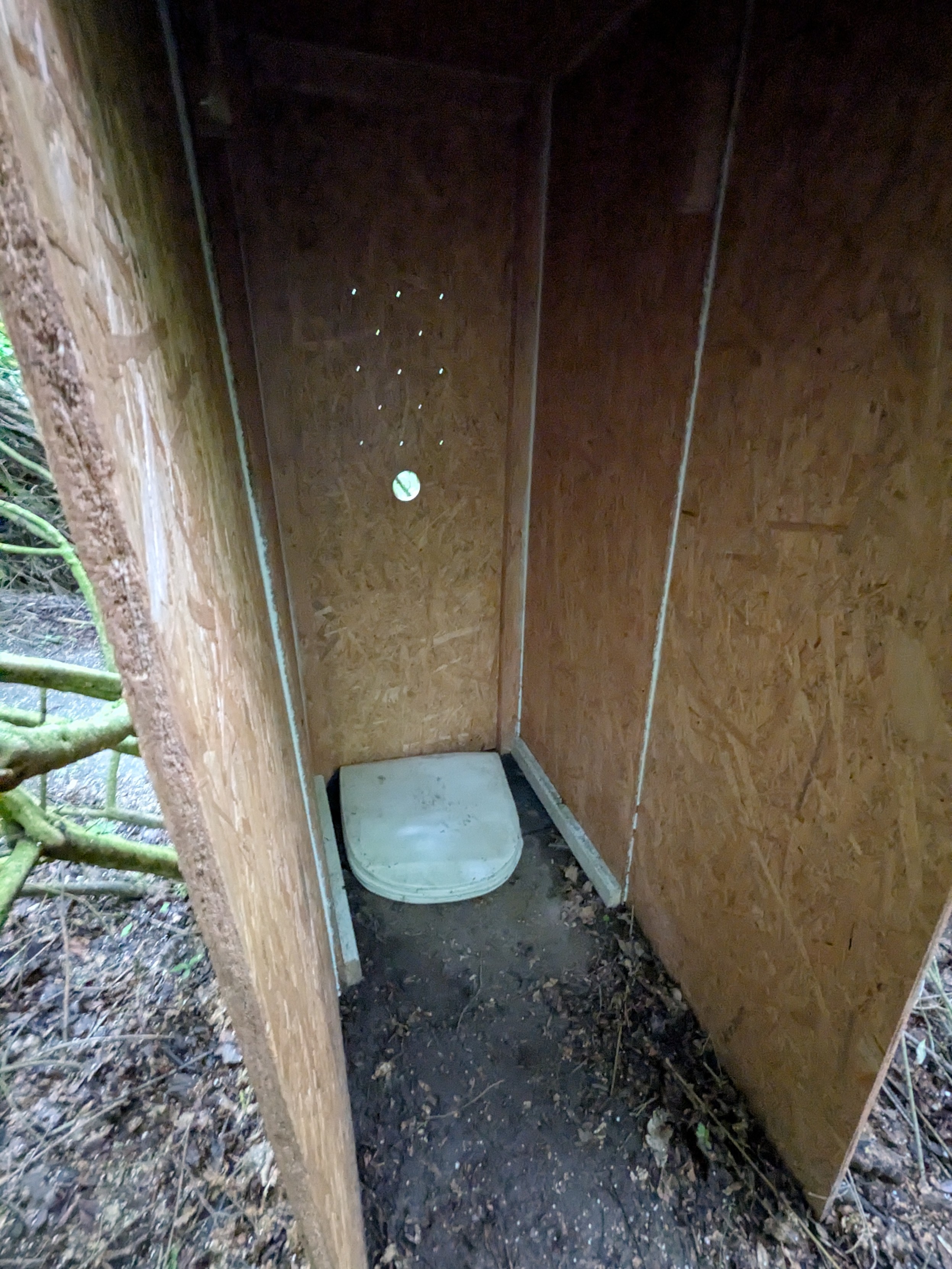 Looking into a simple glory hole booth made from OSB sheets. It has a roof, and the booth is open to one side for easy entrance. The wall opposite to the entrance has a round hole in it, large enough to put a penis through, as well as a couple of small holes in a staggered grid above it, I guess to allow talking through the wall. A chair cushion is lying on the floor to make it more comfortable for someone to kneel in the booth. The floor inside and outside the booth is normal dirt as you would expect. Looking into a simple glory hole booth made from OSB sheets. It has a roof, and the booth is open to one side for easy entrance. The wall opposite to the entrance has a round hole in it, large enough to put a penis through, as well as a couple of small holes in a staggered grid above it, I guess to allow talking through the wall. A chair cushion is lying on the floor to make it more comfortable for someone to kneel in the booth. The floor inside and outside the booth is normal dirt as you would expect.