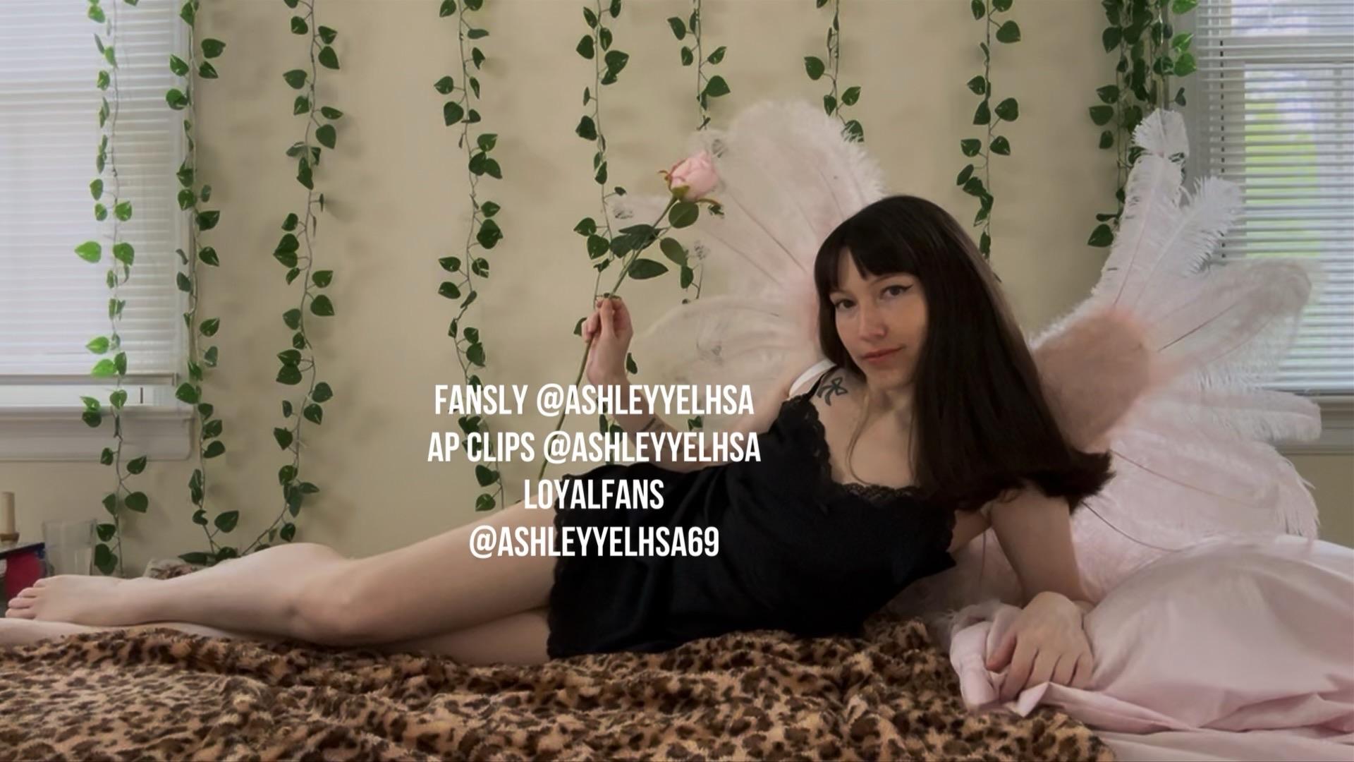 Photo of me, petite white femme with long dark brown hair, laying on my side on a bed, one arm propping me up and my other hand holding a rose, a smirk on my face, wearing a black slip and pink feather wings
