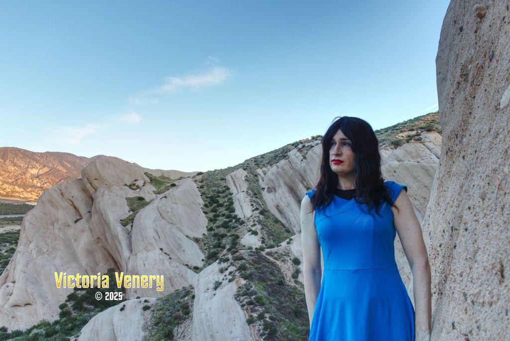 A crossdresser in a blue Starfleet uniform stands in front of a rock formation