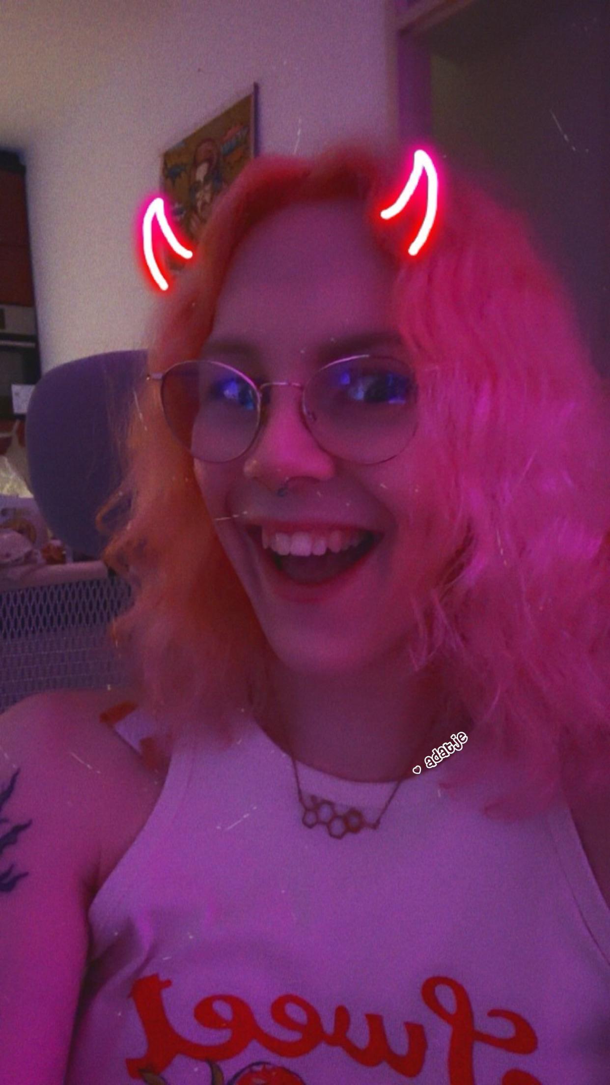 A person with wavy, pink hair wearing glasses and a white tank top is smiling and posing playfully. They have animated devil horns on their head and there is a colorful, softly lit background. The atmosphere is cheerful and vibrant.